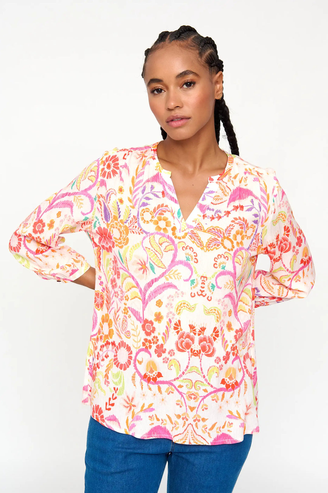Model presents 'Adelaida' style blouse with bright floral print in pink, orange, and yellow, long sleeves, and V-neckline, paired with dark blue jeans.