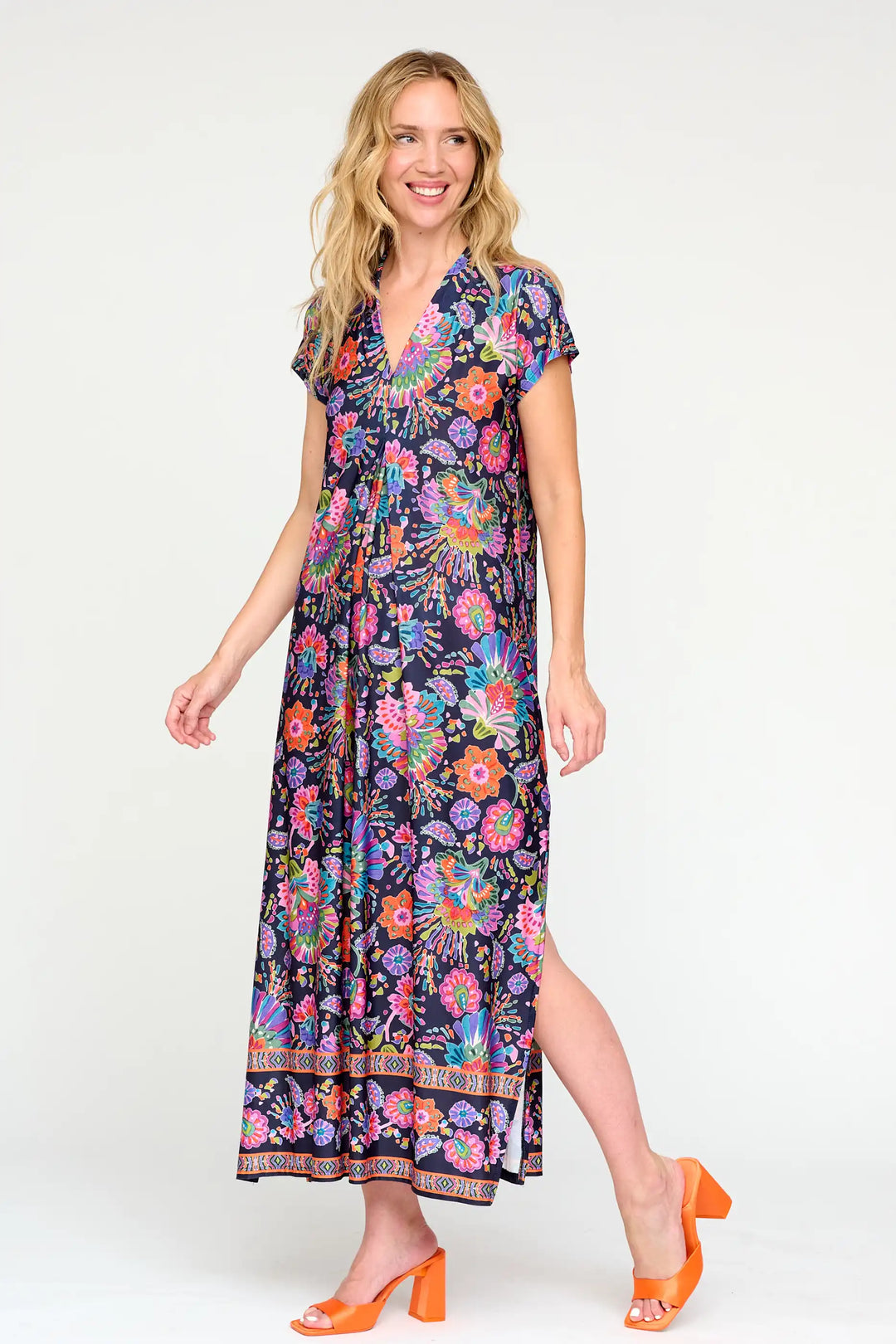 Beaming model in 'Aleli' style navy maxi dress with vibrant pink, purple, and orange floral print, V-neck, short sleeves, and tiered skirt, complemented by bright orange block heel sandals.