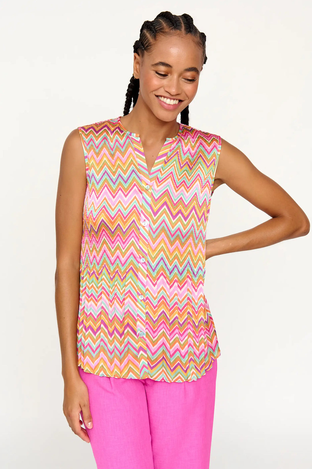 A model with a cheerful demeanor wearing the 'Ambrosia' style sleeveless blouse, featuring a bright, multicolored zigzag pattern, with a collar and front button placket, paired with vibrant pink trousers.
