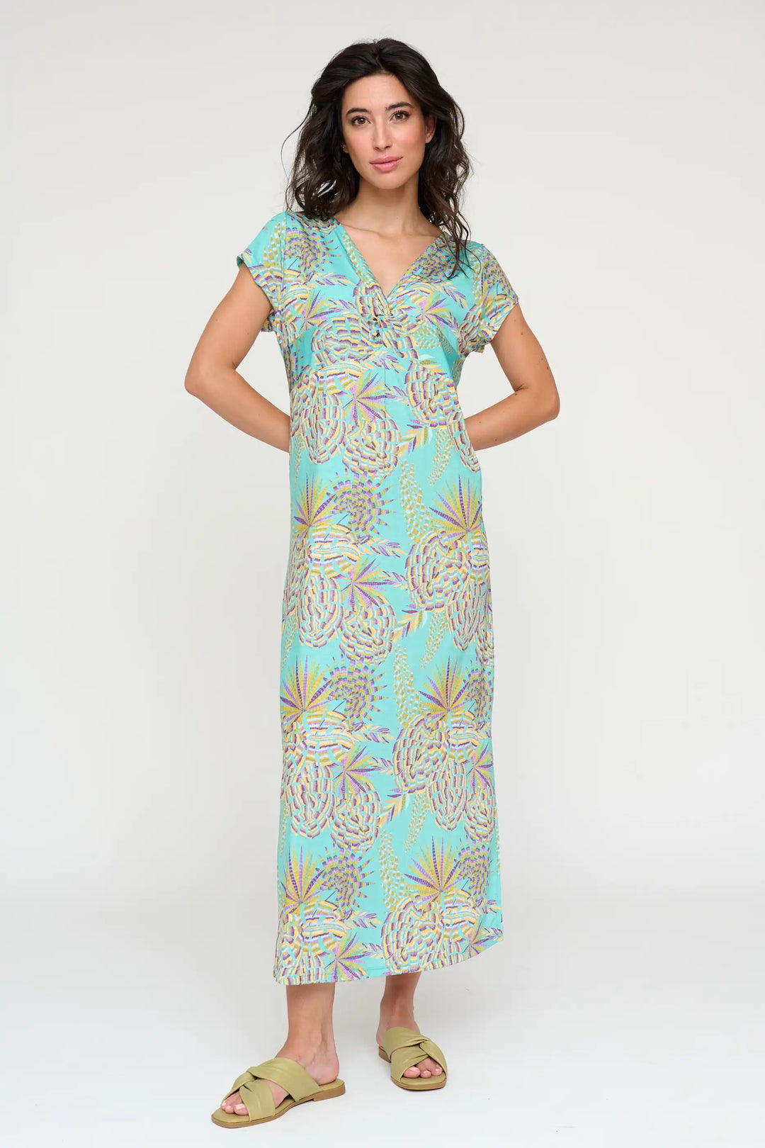 Confident model wearing the 'Amelina' style maxi dress with a tropical palm print in aqua, lavender, and gold, featuring a V-neckline and cap sleeves, paired with olive green slide sandals.