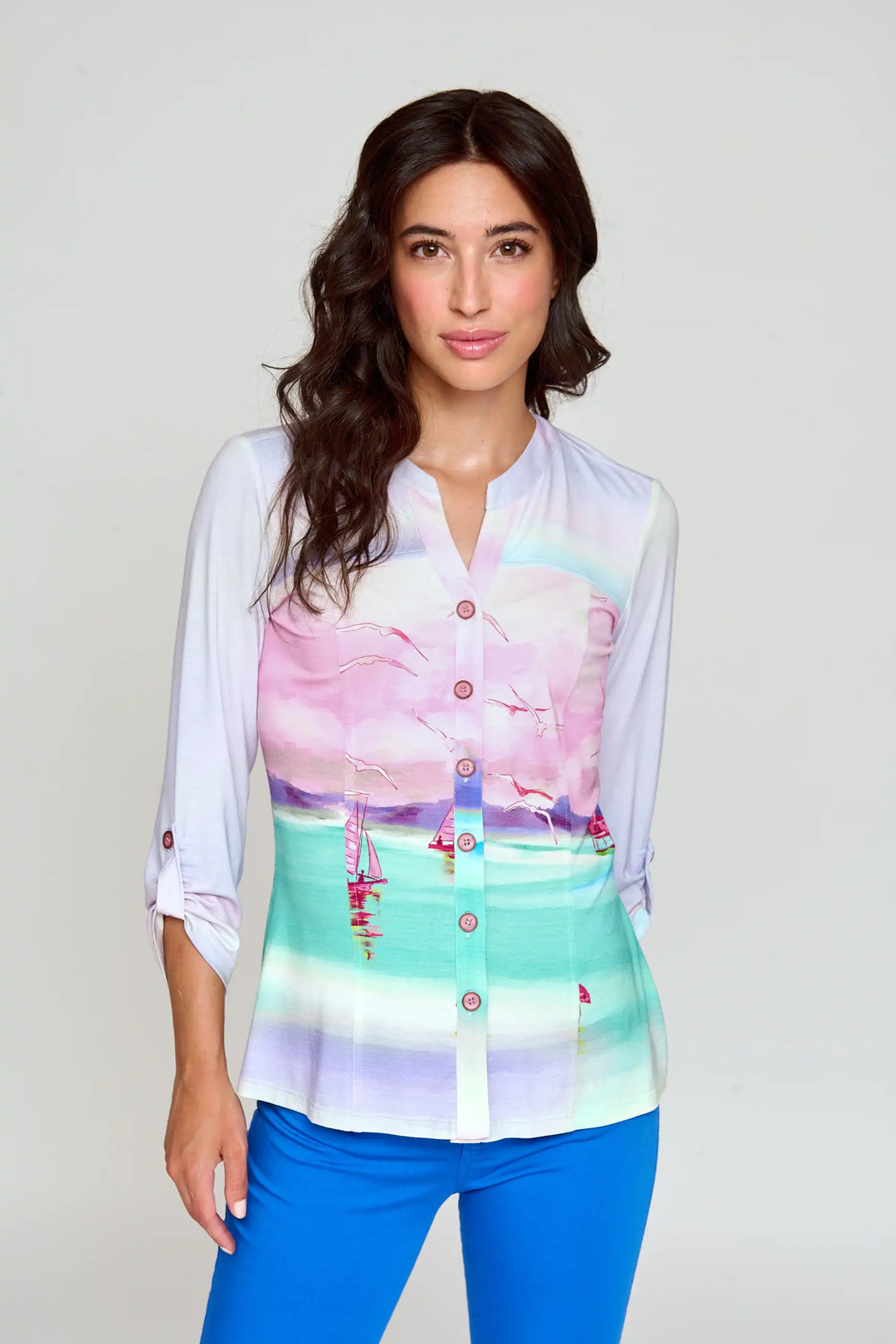 Model wearing the 'Beatrisa' style blouse with a serene seascape print in pastel pink and aqua, long sleeves with buttoned tabs, and a button-up front, paired with bold blue trousers.