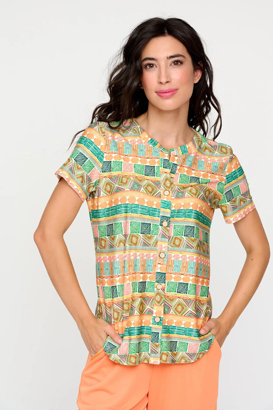 Model wearing the 'Bluma' style blouse, featuring a geometric art deco-inspired pattern in apricot and mint green, with short sleeves and a buttoned front, complemented by peach-coloured trousers.