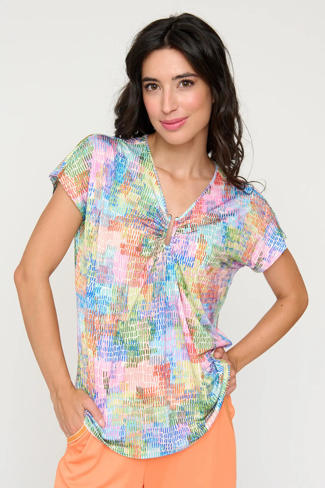 Model wearing the 'Diega' style blouse with a bright, multi-coloured abstract print, cap sleeves, and a V-neckline, tucked into peach-coloured trousers.