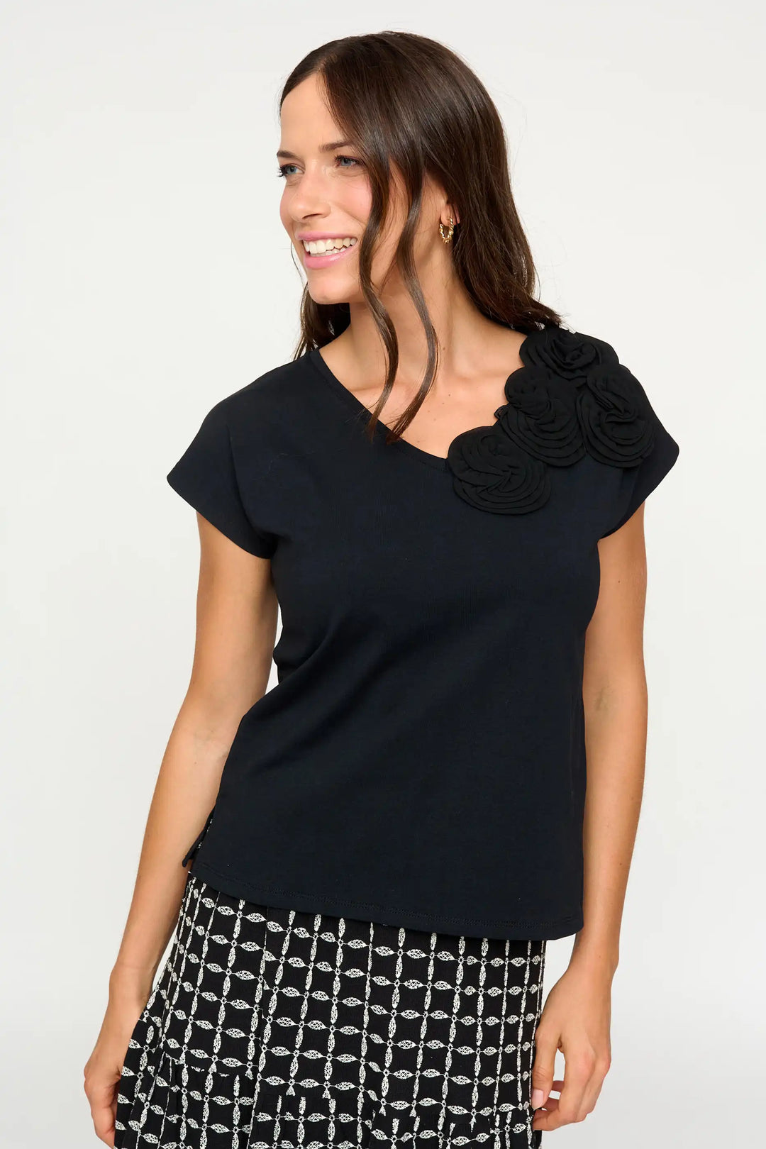 Smiling model in the 'Hinojales' style black top with distinctive floral appliqué on one shoulder, paired with a patterned skirt.
