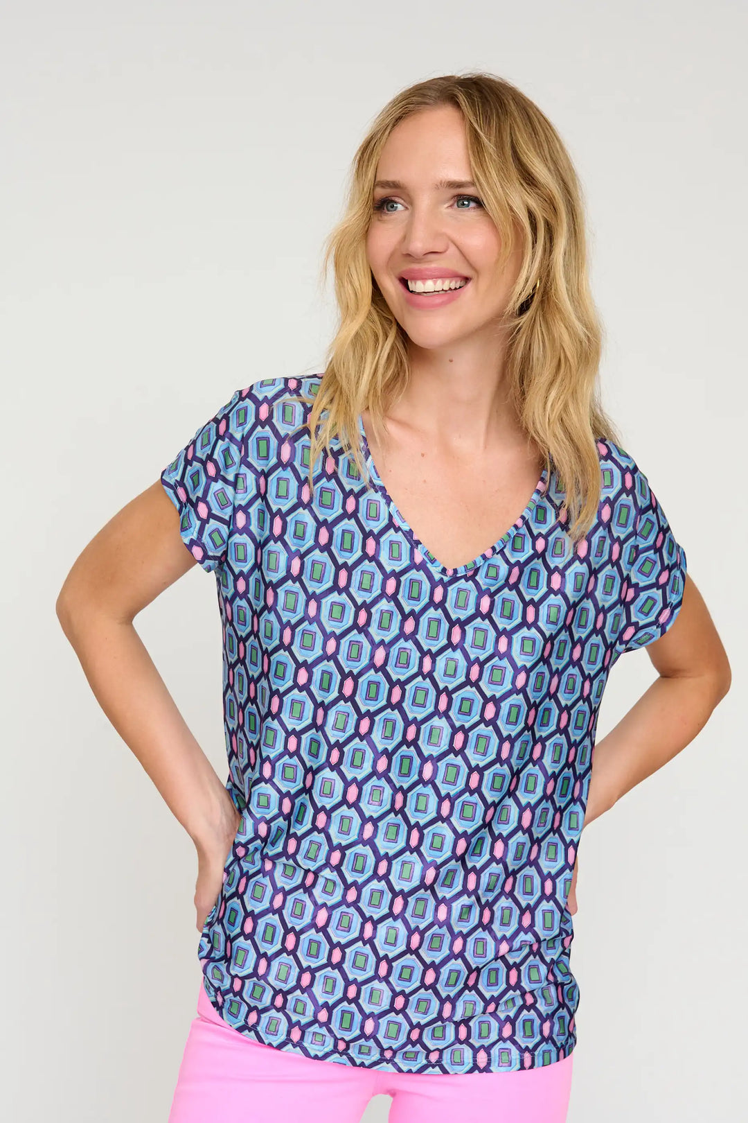 Model wearing the 'Saberina24' style blouse, with a blue and pink geometric print, V-neck, and short sleeves, complemented by pink trousers.