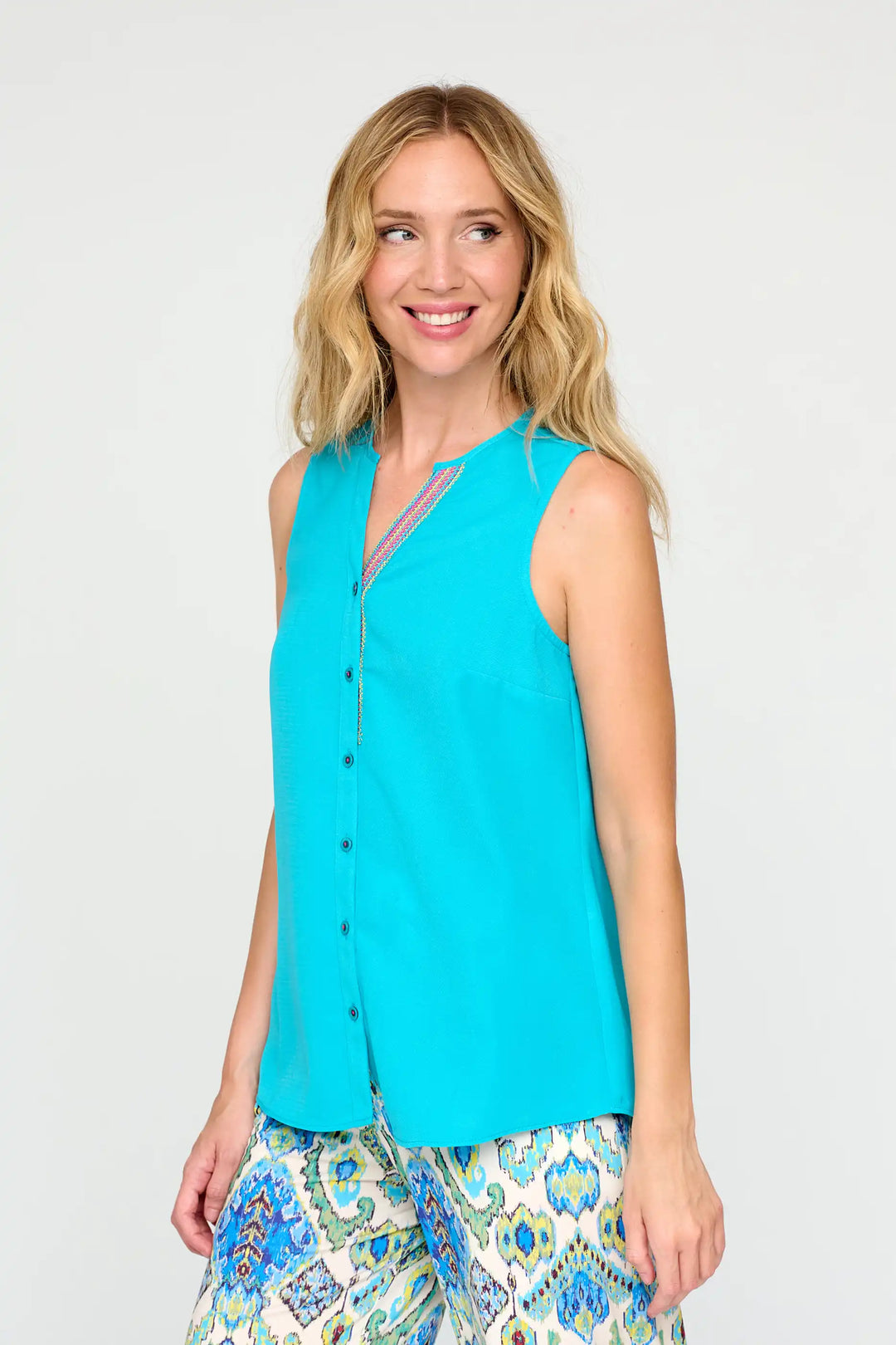A model in the 'Walda24' style top, featuring a bright turquoise colour, sleeveless design, and button-down front, with a touch of multi-coloured trim along the V-neckline.