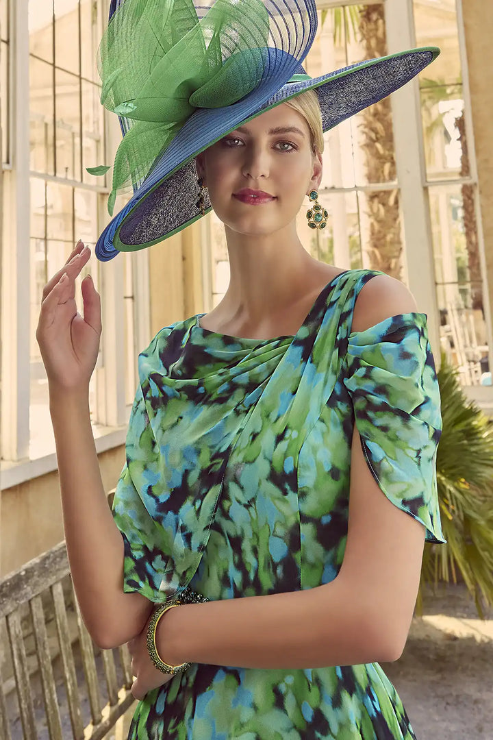 Close-up of woman wearing an Invitations by Veni 36028 midi dress with an abstract green, turquoise, and black print, featuring a scoop neckline, dramatic off-the-shoulder cape, delicate glass bead belt, and a flowing pleated skirt.