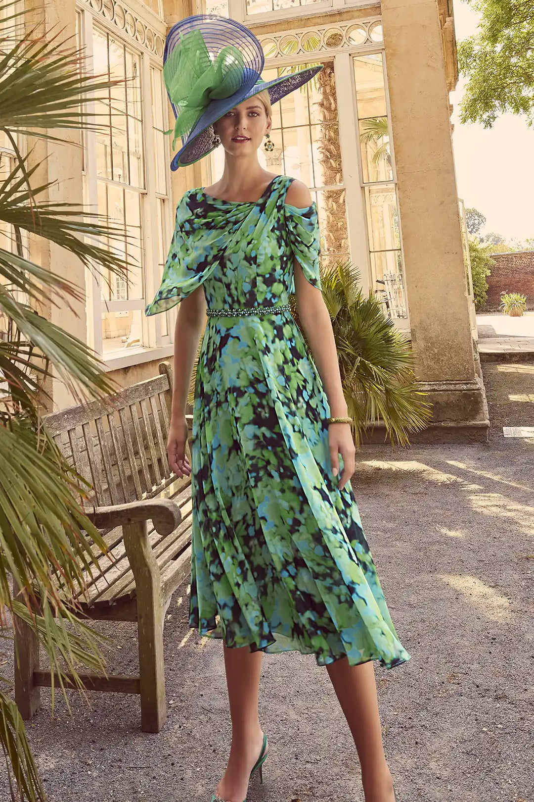 Woman wearing an Invitations by Veni 36028 midi dress with an abstract green, turquoise, and black print, featuring a scoop neckline, dramatic off-the-shoulder cape, delicate glass bead belt, and a flowing pleated skirt.