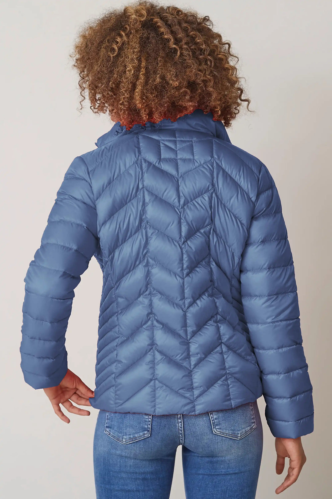 Back view of a woman wearing a slate blue quilted jacket with a high neckline and zip front, over a light blue shirt, paired with denim jeans, style 0124-2040-62-55