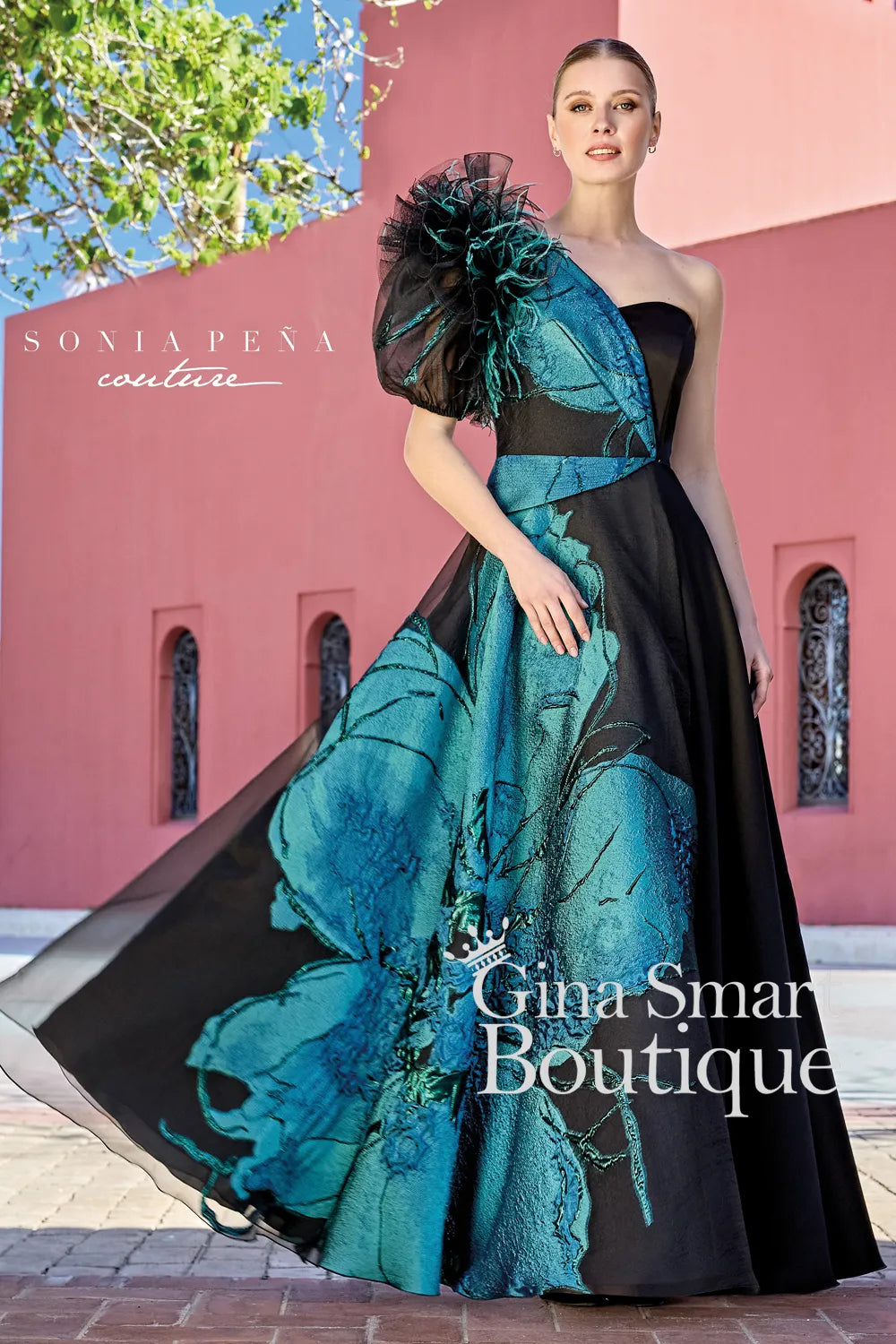 A woman modelling a Sonia Peña Couture evening gown. The dress is a one-shoulder black A-line gown featuring a large metallic teal floral print, a dramatic ruffled puff sleeve with feather accents, and a floor-length skirt.
